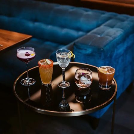 Five colorful craft cocktails — a purple coupe, a chili-rimmed citrus drink, a clear lemon-twist cocktail, a whiskey on the rocks with an ice sphere, and a brown iced cocktail — arranged on a round black glass table in a plush blue velvet cocktail lounge.
