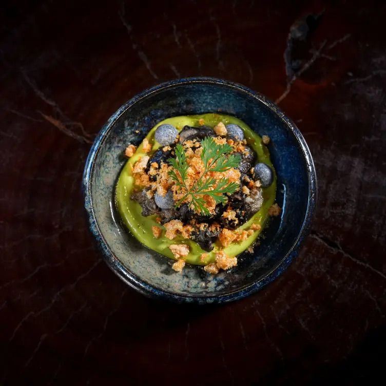 Top-down view of a gourmet blue ceramic bowl with a vibrant green purée ring, blackened crumble center, crunchy golden crumbs, gray discs and a fresh herb sprig on a dark marble table.