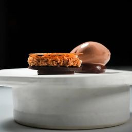 Chocolate plated dessert on a white marble pedestal — dark chocolate discs topped with a crunchy caramelized nut layer and thin brittle, paired with glossy chocolate ice cream and sauce against a black background.