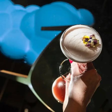 Hand holding a frothy coupe cocktail garnished with a yellow-purple edible pansy, reflected on a glossy black table with blue neon cloud light in the background