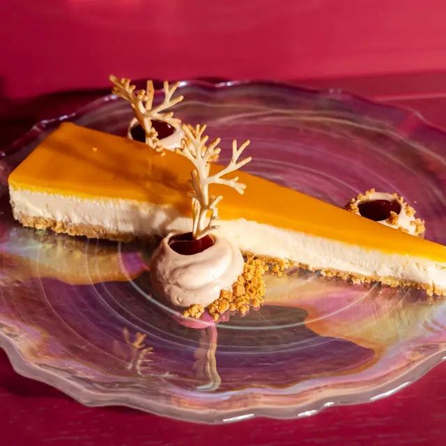 Vibrant mango-topped cheesecake slice with crumb crust, cream dollops with berry centers and white‑chocolate coral garnishes on an iridescent glass plate — elegant plated dessert