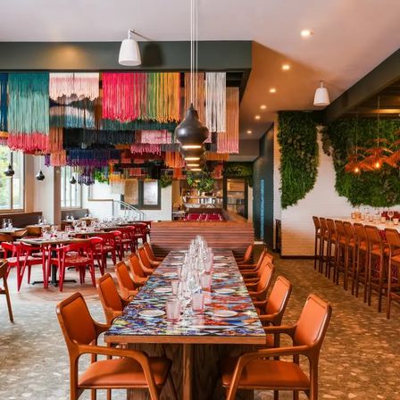 Vibrant modern restaurant interior with a long communal table and orange leather chairs, multicolored hanging yarn ceiling installation, red side seating, a living green wall and a lively bar area.