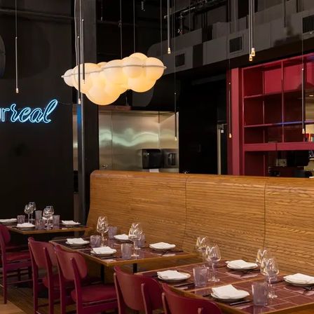 Trendy urban restaurant interior with wooden banquette and maroon chairs, tables set with wine glasses and napkins, glowing cloud-shaped pendant light and neon wall sign, cozy contemporary dining scene.