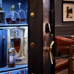LED-lit black safe turned luxury minibar showcasing glass decanters and whiskey bottles on shelves, brass handle and bolts, cozy leather lounge backdrop.