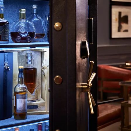 LED-lit black safe turned luxury minibar showcasing glass decanters and whiskey bottles on shelves, brass handle and bolts, cozy leather lounge backdrop.