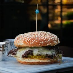 Close-up of a juicy cheeseburger on a sesame seed bun with melty cheese and pickle slices, served on a marble board at a restaurant