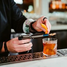 Bartender torching an orange peel to flame citrus oils over an amber cocktail in a lowball glass at a craft cocktail bar