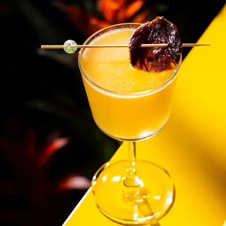 Vibrant mango-yellow cocktail in a stemmed coupe glass, garnished with a skewered dried plum and decorative bead, photographed on a bright yellow surface for a tropical bar-style drink