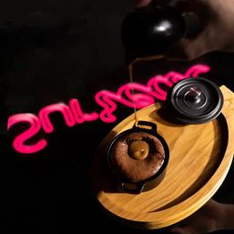 Molten chocolate cake in a mini cast-iron skillet with a gooey caramel center, caramel being drizzled from a small pot onto the dessert on a wooden serving board, framed by a neon pink sign reflection — neon-lit cafe dessert shot.