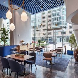 Modern glass-front downtown café with blue slatted ceiling, teardrop pendant lights, plush chairs, potted plants and city street view