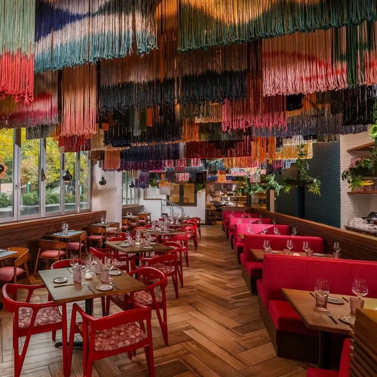 Contemporary restaurant interior featuring a vibrant multicolored fringe canopy overhead, red banquettes and chairs, wooden herringbone floor, set dining tables and large windows with greenery outside