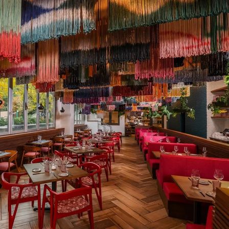 Contemporary restaurant interior featuring a vibrant multicolored fringe canopy overhead, red banquettes and chairs, wooden herringbone floor, set dining tables and large windows with greenery outside
