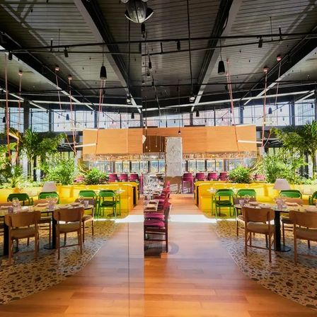 Open‑concept urban restaurant interior with high industrial ceiling and floor‑to‑ceiling windows, wooden central walkway, colorful seating (yellow booths, green chairs, magenta accents), lush indoor plants and bright natural light.