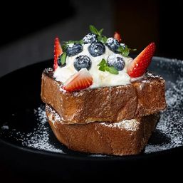 Stacked brioche French toast crowned with whipped cream, fresh blueberries, strawberry slices, mint leaves and a dusting of powdered sugar on a black plate