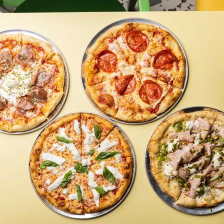Overhead view of four artisan pizzas on metal pans atop a yellow table: classic pepperoni, margherita with fresh basil and ricotta dollops, prosciutto with creamy cheese, and a sliced turkey-style pizza with green pesto and chopped nuts.