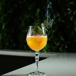 Smoky rosemary-garnished orange cocktail in a faceted crystal stemmed glass with a foamy top and a burning rosemary sprig releasing smoke against a dark green garden backdrop.