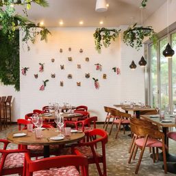 Bright modern café interior with round wooden tables and vibrant red chairs set for dining, white brick wall decorated with small animal masks and pink flowers, hanging greenery and pendant lights, floor-to-ceiling windows showing trees outside.