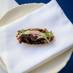 Bite-sized gourmet taco canapé with shredded meat over black bean purée, topped with thin jicama strips, microgreens and a small pink edible flower on a white napkin.