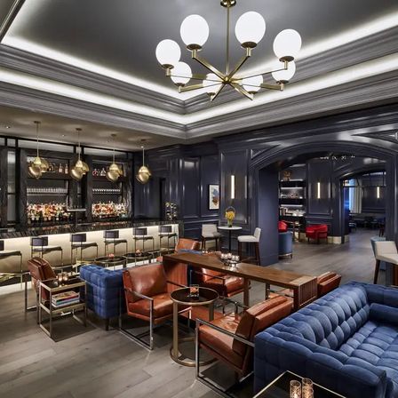 Luxurious hotel bar lounge with navy paneled walls, marble backlit bar, brass pendant and globe chandelier lighting, blue tufted sofas and brown leather chairs