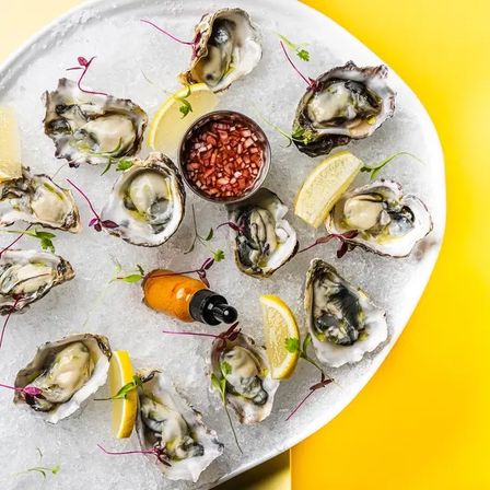 Platter of fresh oysters on the half shell on crushed ice with lemon wedges, mignonette sauce and a hot sauce bottle on a bright yellow background