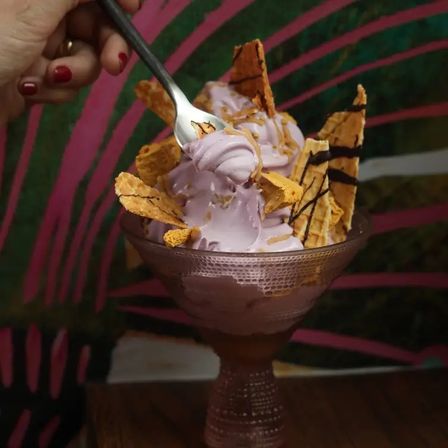 Lavender-purple soft-serve ice cream sundae in a textured glass goblet topped with crispy waffle shards and chocolate drizzle, a spoon scooping a swirl while a hand with red nail polish holds it against a colorful tropical-patterned backdrop.