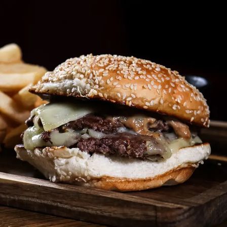 Close-up of a juicy double cheeseburger with melted cheese, savory sauce and sesame-seed bun, served with fries on a wooden board