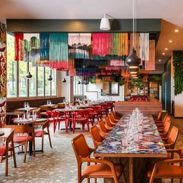 Contemporary restaurant interior with vibrant multicolored fringe ceiling art, long communal table set with glassware, wooden chairs and large windows letting in daylight.