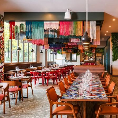 Contemporary restaurant interior with vibrant multicolored fringe ceiling art, long communal table set with glassware, wooden chairs and large windows letting in daylight.