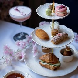 Elegant afternoon tea: three-tier stand of macarons, petits fours, éclair and croissant sandwich with a teacup, pink cocktail and delicate blossoms.
