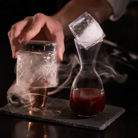 Smoked cocktail presentation: hand lowering a smoke-filled glass cloche over a copper cup next to a carafe of deep red drink topped with a large ice cube.
