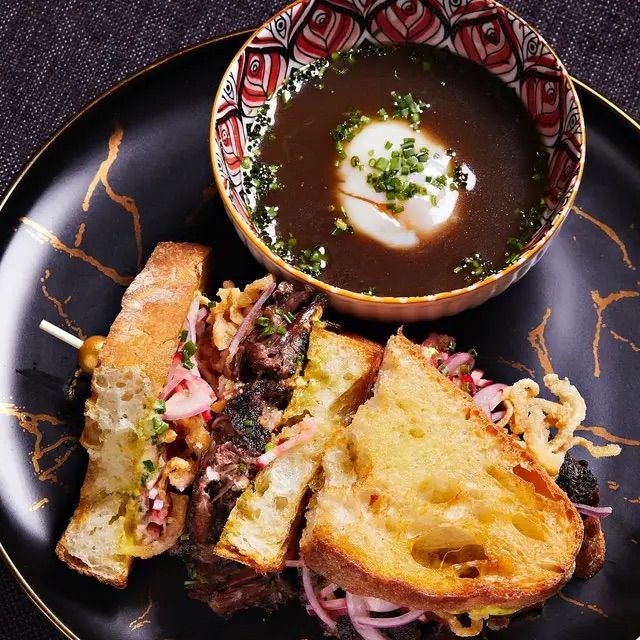 Gourmet grilled beef sandwich on toasted sourdough with pickled onions and crispy bits, served alongside a bowl of rich au jus topped with a soft-poached egg on a black marbled plate.
