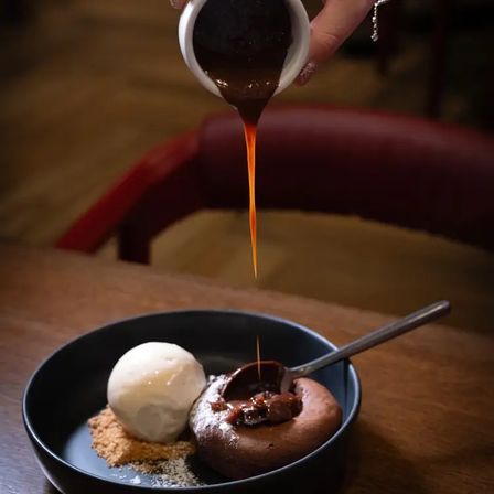 Indulgent molten chocolate lava cake with hot chocolate sauce being poured over it, served with vanilla ice cream and cookie crumbs in a black bowl on a wooden table.