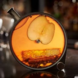 Glowing amber mulled apple cocktail in a round glass carafe on a bar counter, garnished with an apple slice, honeycomb piece, cinnamon stick and whole cloves.