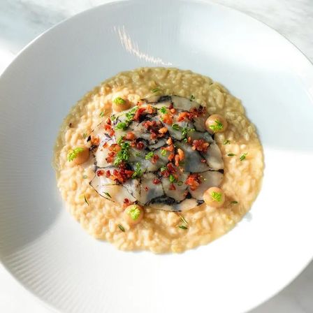 Creamy fine-dining risotto on a white plate topped with thinly sliced seafood carpaccio, crispy cured-meat crumbles, micro-herbs and small sauce dots — elegant restaurant presentation.