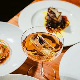 Amber cocktail in a coupe glass with a large clear ice cube, surrounded by gourmet plated dishes on a wooden table — elegant fine-dining drink