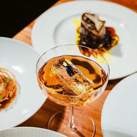 Amber cocktail in a coupe glass with a large clear ice cube, surrounded by gourmet plated dishes on a wooden table — elegant fine-dining drink