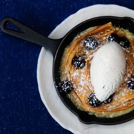 Cast-iron skillet dessert with crispy phyllo nest, powdered sugar, blueberries and a scoop of vanilla ice cream on a white plate over a blue tablecloth