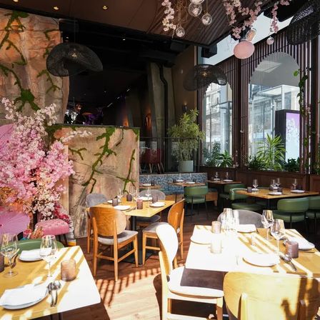 Sunlit modern restaurant dining room with wooden tables and mixed chairs, green banquette seating, pink cherry blossom display, hanging pendant lights, potted plants and large arched windows overlooking the city street.