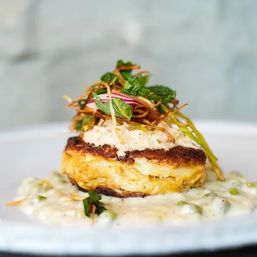 Crispy golden crab cake topped with creamy remoulade, pickled red onion, fried leeks and microgreens, plated on a white dish with herbed cream sauce — gourmet seafood presentation.