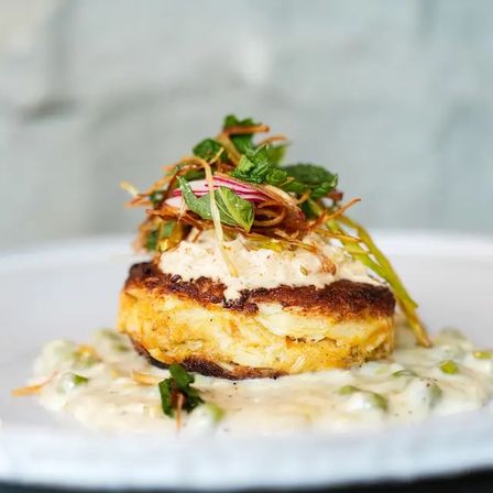 Crispy golden crab cake topped with creamy remoulade, pickled red onion, fried leeks and microgreens, plated on a white dish with herbed cream sauce — gourmet seafood presentation.