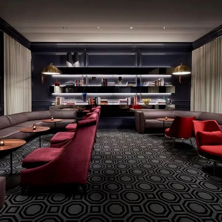 Elegant boutique hotel lounge with dark paneled walls, backlit bookshelves, geometric carpet, plush red velvet chairs, curved gray sofas, brass dome lamps and small wooden tables.
