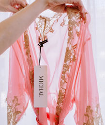 Backlit sheer pink chiffon robe with gold lace trim held up by hands, a white clothing tag dangling from the neckline