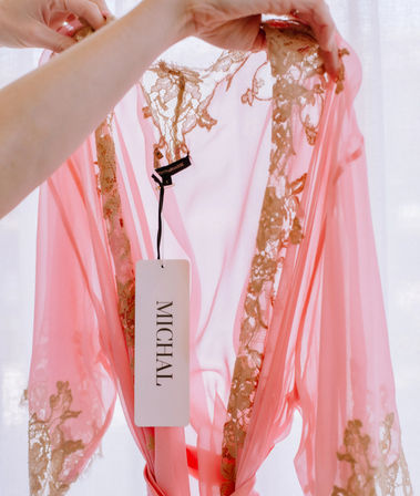 Backlit sheer pink chiffon robe with gold lace trim held up by hands, a white clothing tag dangling from the neckline