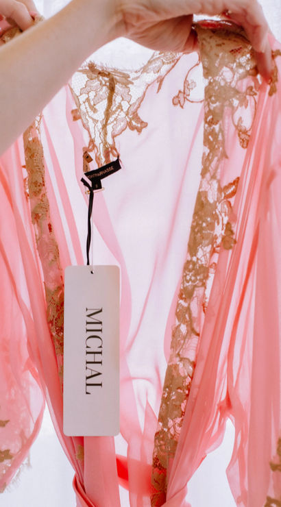 Backlit sheer pink chiffon robe with gold lace trim held up by hands, a white clothing tag dangling from the neckline