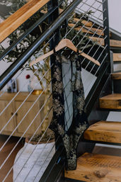 Black lace bodysuit with gold embroidery on a wooden hanger draped over a metal cable railing of a modern wooden-step staircase with a potted plant in a cozy loft interior.