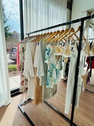 Sunlit boutique clothing rack by a floor-to-ceiling window with wooden hangers holding airy neutral dresses, blue floral tops and gold-lace lingerie over wood floors, storefront visible outside.