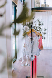 Chic window-lit boutique display of a delicate white lace bodysuit and pink silk robe on wooden hangers, with a mannequin and potted plant in an airy modern showroom with floor-to-ceiling windows.