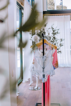 Chic window-lit boutique display of a delicate white lace bodysuit and pink silk robe on wooden hangers, with a mannequin and potted plant in an airy modern showroom with floor-to-ceiling windows.