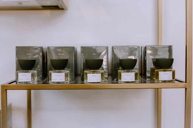 Five luxury perfume bottles with round glass bases and matte black bowl caps lined up on a brass display shelf against a white wall — minimalist boutique fragrance display.