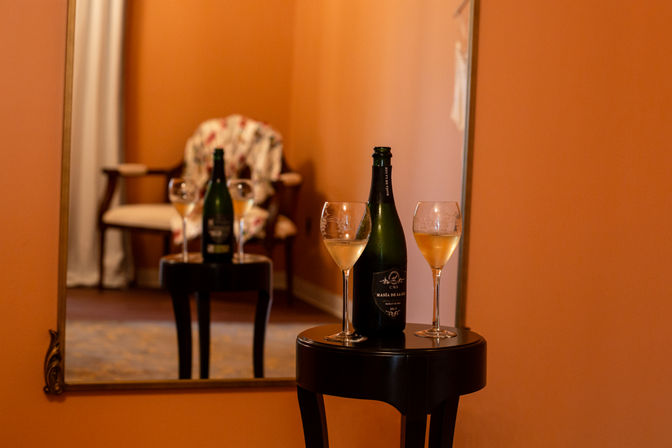 Bottle of sparkling wine and two filled wine glasses on a small black side table in a cozy hotel suite, reflected in a large mirror with a floral armchair and warm orange walls.